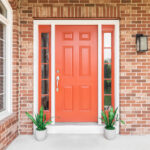 A brick home’s red front door surrounded by plants sitting in front of windows.