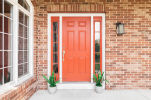 A brick home’s red front door surrounded by plants sitting in front of windows.